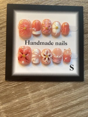 Handgjorda press-on naglar med blommor - Säljer ett set handgjorda press-on naglar i rosa och vitt med 3D-blommor, pärlor och gulddetaljer. Naglarna har en kort, rundad form och dekorerade med olika mönster och små pärlor för en söt och unik look. Perfekt för dig som vill ha något extra till naglarna.