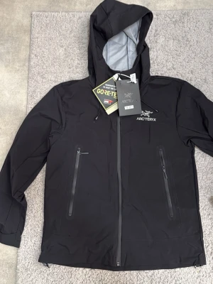 Svart Arc'teryx regnjacka med huva - Snygg svart regnjacka från Arc'teryx med huva och vattentätt GORE-TEX-material. Jackan har två stora dragkedjeförsedda fickor framtill och justerbara ärmslut. Perfekt för regniga dagar och outdoor-äventyr.