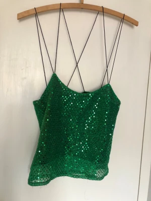Grön paljett-topp med tunna band - Snygg grön topp med glittriga paljetter och tunna svarta axelband som korsas i ryggen. Toppen är ärmlös och croppad, perfekt för dig som vill sticka ut. Materialet känns lätt och skimrande. Snygg till jeans till vardags eller uppklätt till fest.