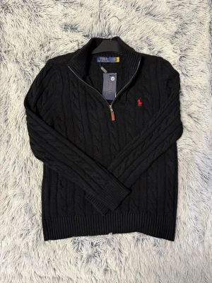 Svart kabelstickad tröja Polo Ralph Lauren - Kabelstickad svart tröja från Polo Ralph Lauren med halv dragkedja och ribbad krage. Tröjan har det klassiska röda Polo-logotypen broderad på bröstet och är tillverkad i mjukt material. Perfekt för lager på lager och stilren look.