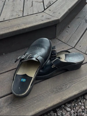 Svarta träskor med spänne - Säljer ett par klassiska svarta träskor i läder med justerbart spänne över foten. De har en robust sula och svensk flagga på innersulan. Perfekta för dig som arbetar.