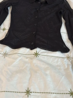 Svart meshskjorta med knappar - Snygg svart meshskjorta med lång ärm och klassisk krage. Skjortan har genomskinligt material och knäppning framtill. Perfekt för att addera en edgy vibe till din outfit.