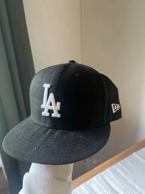 Svart LA New Era keps - Säljer en svart keps från New Era med LA-logga broderad i vitt framtill och MLB-logga bak. Klassisk flat brim och stilren design, perfekt för dig som gillar streetwear och sportig stil. Fram på  rimmen är det typ en böjning som man kan se men inge fel bara ser ut så.