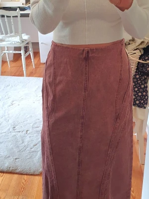 Långkjol i vinröd denim - Säljer en långkjol i mörkrosa denim med markerade sömmar och dragkedja bak. Kjolen har en rak passform och är perfekt för dig som gillar en stilren och trendig look. Passar till både stickade tröjor och crop tops.