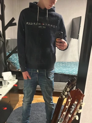 Mörkblå hoodie Tommy Hilfiger - Snygg mörkblå hoodie från Tommy Hilfiger med vit logga och text på bröstet. Klassisk känguruficka framtill och vita dragsnören i huvan. Perfekt för en avslappnad och trendig stil. Mjuk bomullskänsla och ribbade muddar vid ärmslut och nederkant.