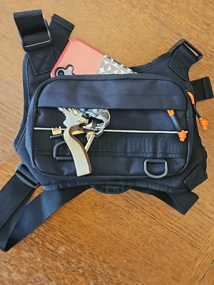 Svart taktisk crossbody-väska - Säljer en svart taktisk crossbody-väska med flera fack och justerbara remmar. Väskan har orange detaljer, reflexband och smarta fästen för nycklar eller andra småprylar. Perfekt för dig som vill ha en praktisk och snygg accessoar till vardagen. Perfekt även är mc - motorcykel. Sommarväska / becknarväska