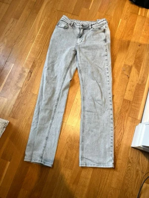 Ljusgrå raka jeans - Säljer ett par ljusgrå jeans med rak passform och klassisk femficksdesign. Jeansen har normal midja och är tillverkade i denim med synliga sömmar. Perfekta för en avslappnad och trendig look.