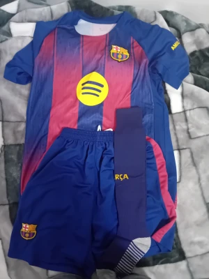 FC Barcelona matchställ med strumpor - Säljer ett komplett FC Barcelona matchställ med tröja, shorts och strumpor. Tröjan har klassiska blå och röda ränder, klubbmärke på bröstet och sponsorlogga. Shortsen är blå med klubbmärke och strumporna är mörkblå med gul text. Perfekt för fotbollsträning eller match.