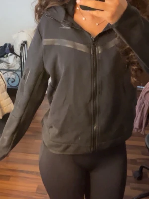 Svart Nike tech - Svart hoodie från Nike med dragkedja framtill och diskreta grå detaljer över bröstet. Hoodien har huva och en ficka på ärmen. Tillverkad i mjukt material som passar perfekt till en sportig eller avslappnad stil.
