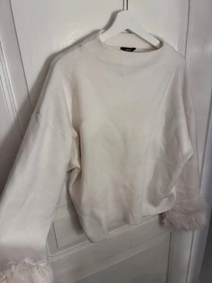 Vit tröja med fuskpälsdetaljer Lindex - Säljer en vit tröja från Lindex med vida ärmar och fluffiga fuskpälsdetaljer vid ärmsluten. Tröjan har en avslappnad passform och rund halsringning. Perfekt för dig som gillar en trendig och mysig stil.