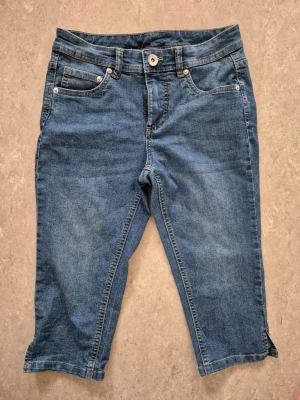 Ankellånga shorts - Säljer ett par blåa ankellånga shorts i klassisk denim med fem fickor och knappgylf. Jeansen har normal midja och raka ben som slutar strax under knät. Perfekta för en avslappnad och cool stil. Materialet är mjukt och stretchigt för extra komfort.