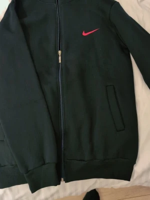 Svart zip hoodie från Nike - Svart hoodie från Nike med dragkedja framtill och två fickor. Klassisk Nike Swoosh i rött broderat på bröstet. Tillverkad i mjukt sweatshirtmaterial, ribbade muddar vid ärmslut och nederkant. Perfekt för en sportig och avslappnad stil. Helt ny passar s & m