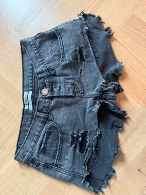 Grå slitna jeansshorts - Säljer ett par grå jeansshorts med, fransiga kanter och slitningar framtill och baktill. Shortsen har fem fickor, knappgylf och en cool, lite trashad look. Perfekta för sommaren, storlek 26.