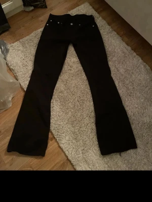 Svarta bootcut jeans vintage - Säljer ett par svarta bootcut jeans med klassisk femficksmodell och silverfärgade nitar. Jeansen har låg midja och vida ben, perfekta för en trendig och avslappnad look. Materialet är kraftigt denim i bomull. Passar dig som gillar retrostil!