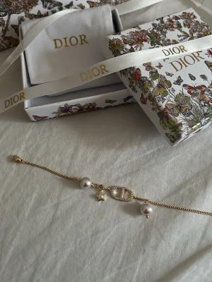 Armband med pärlor och stjärna - Lyxigt armband i guldton med två vita pärlor, en liten stjärnberlock och ett gnistrande CD-emblem i mitten. Armbandet har en tunn kedja och detaljer som ger en elegant och trendig vibe. Perfekt accessoar för dig som vill sticka ut lite extra. Utan paketering är det 350kr men med paketering 550kr.