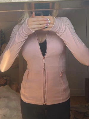 Ljusrosa lululemon träningsjacka med dragkedja - Snygg ljusrosa träningsjacka från lululemon som är väldigt trendig just nu med figurnära passform och hel dragkedja framtill. Jackan har långa ärmar med tumhål, två fickor med dragkedja. Tillverkad i ett stretchigt och mjukt material som passar perfekt till gymmet eller löprundan. Den har roseguld detaljer på dragkedjan!