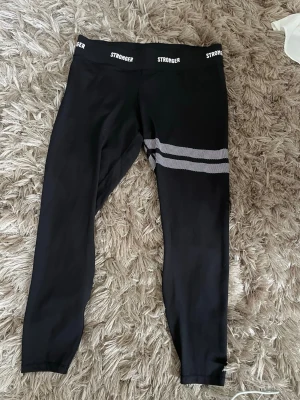 Svarta tights från Stronger - Snygga svarta träningstights från Stronger med vita detaljer i form av två ränder på ena benet. Hög midja med logotyp runt linningen. Tillverkade av 75% återvunnen polyester, perfekta för gymmet eller löprundan.