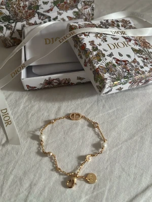 Armband med pärlor och guldton - Lyxigt armband i guldtonat metall med vita pärlor och detaljerad CD-logga med små stenar. Kedjan har en rund och smidig karbinhake. Kommer i exklusiv presentask med pärligt mönster. Utan box, dustbag och paketering är det 350kr och med paketering 550kr