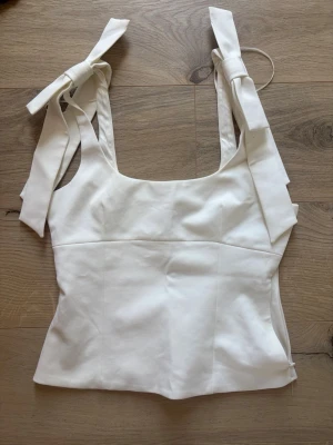 Vit topp med knytband från Zara Xs - Supersnygg vit topp från Zara med breda axelband som knyts till rosetter. Toppen har en fyrkantig ringning och är figurnära i modellen. Perfekt att styla med jeans eller kjol för en fräsch look.