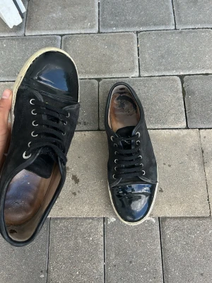 Lanvins  - Säljer ett par svarta sneakers med snygga lackdetaljer på tån och vita sulor. Skorna har svarta skosnören och metallfärgade öljetter. Insidan är brun och materialet är en mix av mocka och lack. Perfekta för dig som gillar en stilren men ändå unik look.