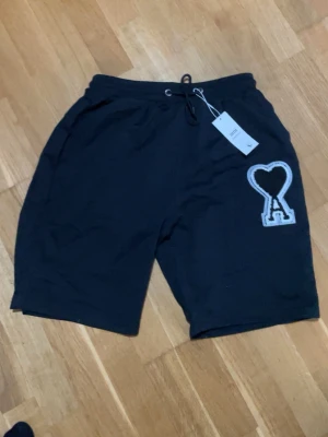 Svarta shorts från Ami Paris med hjärtdetalj - Svarta shorts från Ami Paris med vit broderad hjärtdetalj på ena benet. Modellen har snörning i midjan och avslappnad passform. Perfekta för en stilren och bekväm look. Materialet är mjukt och känns skönt mot huden. Komm med bud vid snabb affär 
