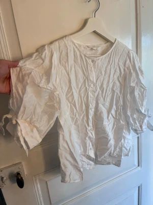 Vit blus med knappar från H&M - Säljer en vit blus från H&M med rund halsringning och knappar framtill. Blusen har trekvartsärm med knytband vid ärmsluten och är tillverkad i ett tunt, luftigt bomullstyg. Perfekt för en avslappnad och trendig look.
