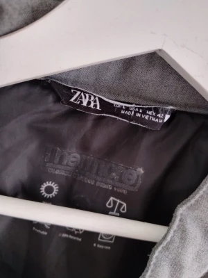 Grå dunväst från Zara - Snygg grå dunväst från Zara med ståkrage och svarta tryckknappar. Västen har två framfickor med dragkedja och är fodrad med Thermic material för extra värme. Perfekt för lager på lager och enkel att matcha med olika outfits.