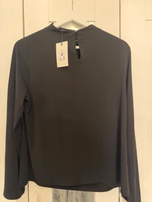 Svart långärmad blus från Bikbok  - Stilren svart blus från Bikbok  med lång ärm och diskret knapp i nacken. Blusen har en rak passform och är tillverkad i ett mjukt, slätt material som ger en elegant look. Perfekt till både jeans och kjol för en trendig outfit. Blusen är aldrig använd.