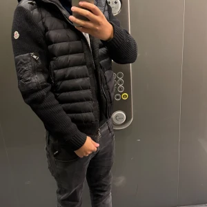 Svart Cardigan från Moncler - Säljer en svart pufferjacka från Moncler med stickade ärmar och huva. Jackan har dragkedja framtill, ficka med dragkedja på ärmen och Moncler-logga på axeln. Materialmix av polyester och stickat ger en snygg och modern look.