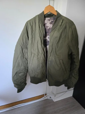 Olivgrön bomberjacka med camofoder - Snygg olivgrön bomberjacka med ribbade muddar och dragkedja framtill. Insidan har ett coolt camouflagemönstrat foder som ger extra edge. Två snedställda fickor på framsidan. Perfekt för dig som vill ha en klassisk streetstil. Går ha åt båda håll