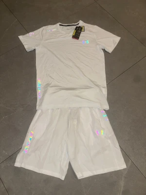 Vitt set från Under Armour - Säljer ett vitt set från Under Armour med t-shirt och shorts. Setet har tryckta loggor och detaljer i svart samt holografiska effekter på vissa delar. Materialet är lätt och andas, perfekt för en sportig look. Passar dig som gillar stilrena och moderna träningskläder.