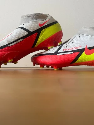 Nike Phantom GT fotbollsskor - Snygga Nike Phantom GT fotbollsskor i vitt, rött och gult med svarta detaljer. Skorna har en strumpliknande passform och är tillverkade i syntetmaterial för bra bollkontroll. Perfekta för dig som vill sticka ut på planen med grym design och funktion. Utan skosnören ( kan lägga till själv ) pris kan diskuteras.