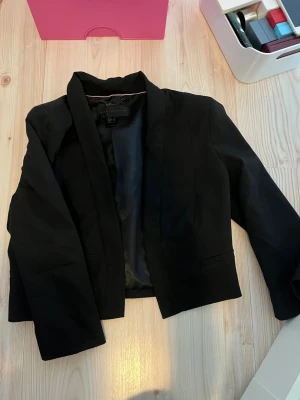 Svart kavaj från MNG Collection - Stilren svart kavaj från MNG Collection med klassisk krage och öppen front. Kortare modell som ger en modern look. Fodrad insida med diskret rosa kantband. Perfekt att matcha till olika outfits för en trendig stil.