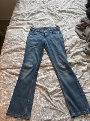 Blå bootcut jeans från Lee - Säljer ett par klassiska blå bootcut jeans från Lee. Jeansen har fem fickor, normal midja och är tillverkade i slitstark denim. Snyggt utställda ben och Lee-lapp bak i midjan. Perfekta för en avslappnad och trendig look. Är ej stretchiga och rätt tunnt material.