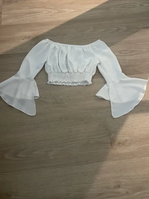 Vit offshoulder croppad blus - Trendig vit offshoulder blus med vida trumpetärmar och smockad midja. Croppad modell som ger en snygg siluett och passar perfekt till höga jeans eller kjol. Luftigt material som känns lätt och bekvämt.