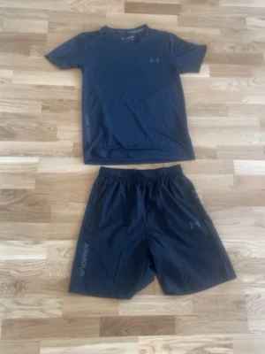 Svart träningsset från Under Armour - Snyggt svart träningsset från Under Armour med t-shirt och shorts. Setet är gjort i ett lätt och snabbtorkande material, perfekt för gymmet eller löpning. Diskreta loggor på både tröja och shorts samt elastisk midja på shortsen för skön passform.