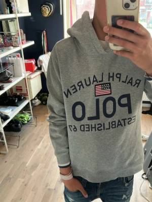 Grå hoodie från Ralph Lauren Polo - Säljer en grå hoodie från Ralph Lauren Polo med stor logotyp och amerikansk flagga på bröstet. Hoodien har långärmad design, ribbade muddar och klassisk huva. Materialet är mjukt och bekvämt, perfekt för en avslappnad look.