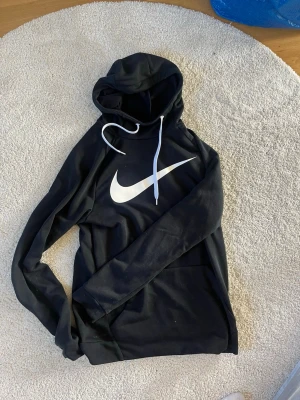 Svart Nike hoodie med vit logga - Säljer en svart hoodie från Nike med stor vit Swoosh-logga på bröstet och vita dragsnören i huvan. Tillverkad i Dri-FIT-material som andas och känns skönt mot huden. Perfekt för dig som gillar sportig och clean stil.