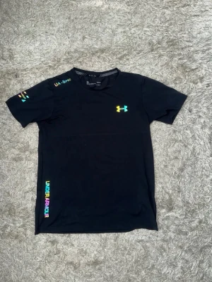 Svart t-shirt från Under Armour - Snygg svart t-shirt från Under Armour med färgglada loggor på bröstet, ärmen och längs sidan. Tillverkad i ett lätt och snabbtorkande funktionsmaterial, perfekt för träning eller sportaktiviteter. Rund halsringning och korta ärmar.