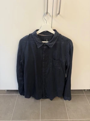 Mörkblå linneskjorta från Osklen, stl L - Snygg mörkblå skjorta från Osklen i linne. Klassisk krage, bröstficka och knäppning framtill. Perfekt för dig som gillar en enkel men stilren look. Skjortan har långa ärmar och en avslappnad passform.