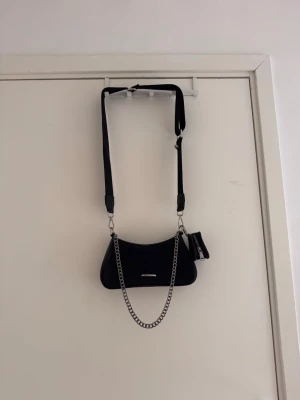 Svart axelväska med kedja - Snygg svart axelväska med justerbart band och silverfärgad kedjedetalj. Väskan har en minimalistisk design och ett litet extra fack med dragkedja. Tror den är ifrån madlady. Använt typ 2 gånger 