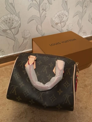 Louis Vuitton Speedy Monogram väska - Säljer en klassisk Louis Vuitton Speedy handväska i brunt monogrammönstrat canvas med ljusa läderdetaljer och guldfärgade metalldelar. Väskan har dragkedja upptill och två handtag. Ikonisk design som passar till många stilar.