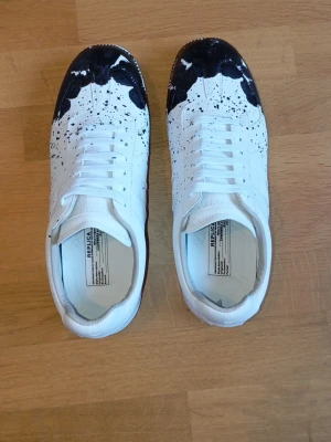 Maison Margiela vita sneakers med svart färg stänk - Unika vita sneakers från Maison Margiela i läder med svart färgstänk och konstnärlig design på tån. Klassisk låg modell med vita skosnören och diskret sömnad. Perfekta för dig som vill sticka ut med en kreativ och trendig look.