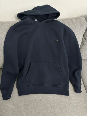 Mörkblå hoodie från Hugo Boss - Snygg mörkblå hoodie från Hugo Boss med broderad logga på bröstet och en gul detalj under. Klassisk känguruficka framtill och ribbade muddar. Perfekt för en chill och stilren look.