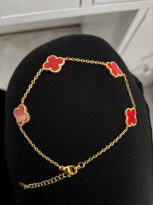 Guldarmband med röda klöver - Snyggt armband i guldton med fyra röda klöverformade hängen. Kedjan är tunn och smidig med ett klassiskt lås. Klöverhänget har en guldfärgad kant som ger en exklusiv känsla. Perfekt accessoar för att lyfta din outfit.