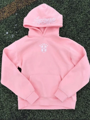 Rosa hoodie från Avenue med tryck - Säljer en ljusrosa hoodie från Avenue med broderad logga och text på huvan. Hoodien har en klassisk känguruficka och ribbade muddar. Perfekt för dig som gillar streetwear och vill ha något bekvämt och snyggt. DE FINNS I ALLA STORLEKAR!!!