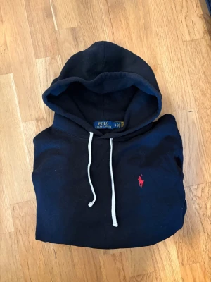 Svart Ralph Lauren hoodie - Klassisk svart hoodie från Polo Ralph Lauren med vit dragsko i huvan och den ikoniska röda loggan broderad på bröstet. Tillverkad i mjukt bomullsmaterial och har en avslappnad passform. Perfekt för en chill och stilren look.