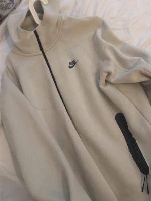 Beige Nike hoodie med dragkedja - Snygg beige hoodie från Nike med svart dragkedja och svarta detaljer vid fickorna. Hoodien har en hög krage, huva och Nike-logga på bröstet. Tillverkad i mjukt material som känns skönt mot huden. Perfekt för en avslappnad streetstyle-look.