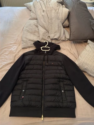 Marinblå cardigan från Tommy Hilfiger - Snygg marinblå pufferjacka från Tommy Hilfiger med huva och dragkedja framtill. Jackan har quiltad kropp, släta ärmar och två fickor med knappstängning. Liten logga på bröstet och vid fickan. Perfekt för kalla dagar.