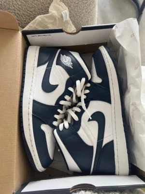 Air Jordan 1 Mid Obsidian/Vit - Säljer ett par Air Jordan 1 Mid i färgen Obsidian och vit. Klassisk siluett med vita läderpaneler och mörkblå detaljer, vita skosnören och den ikoniska Air Jordan-loggan på sidan. Perfekta för dig som gillar streetwear och sneakers med retrovibe.